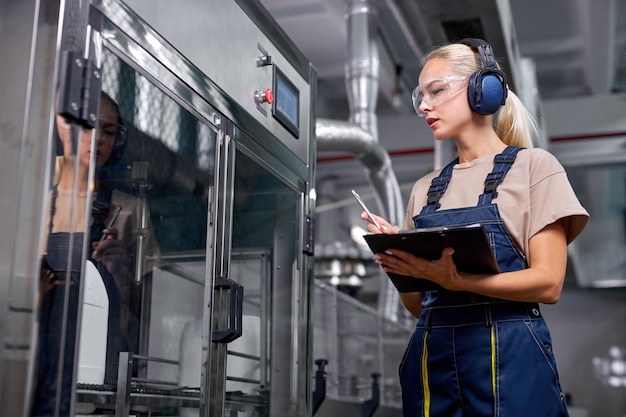 Female factory worker hold quality control at work, take notes into folder-tablet, writing information about robotic equipment. worker in protective uniform working in medical supplies research