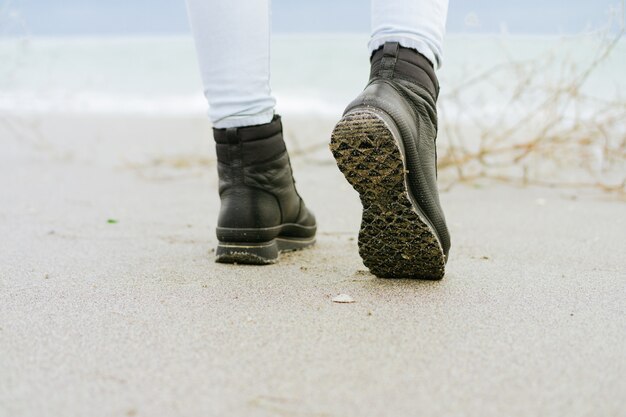 Female feet in blue jeans and black winter boots standing on the beach against the sea