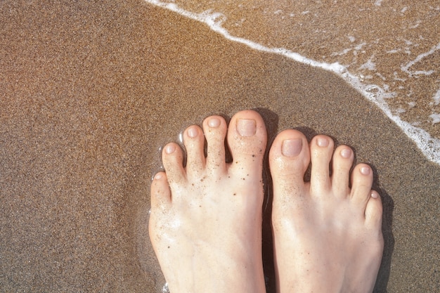 Female feet with beige nail design. Glitter beige nail polish pedicure on ocean beach sand. Summer vacation pedicure.