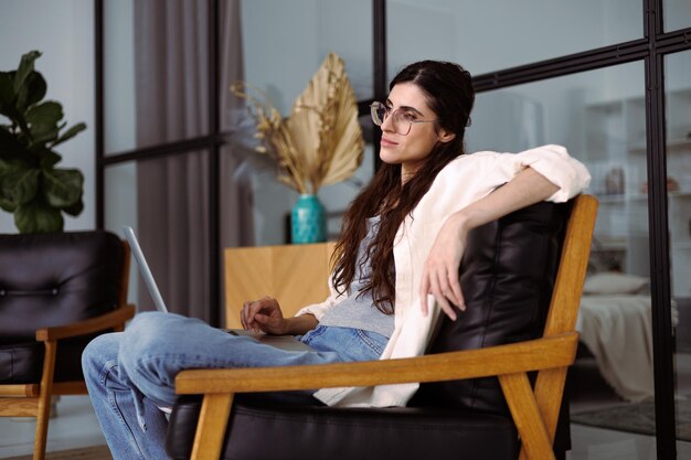 Photo female freelancer in eyeglasses holding laptop while sitting on armchair at home