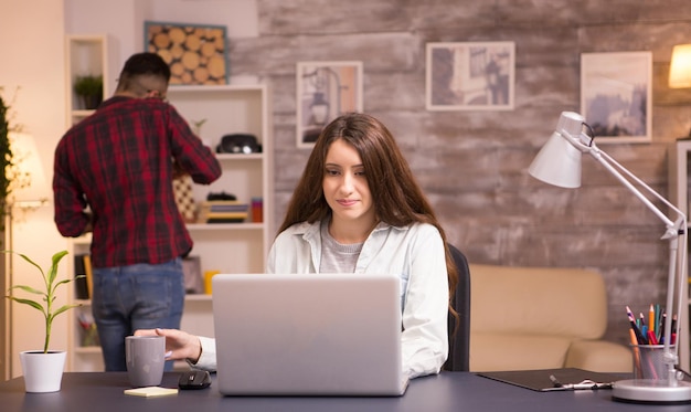 Female freelancer reaching for coffee while working on laptop in living room . Boyfriend talking on his phone in the background.