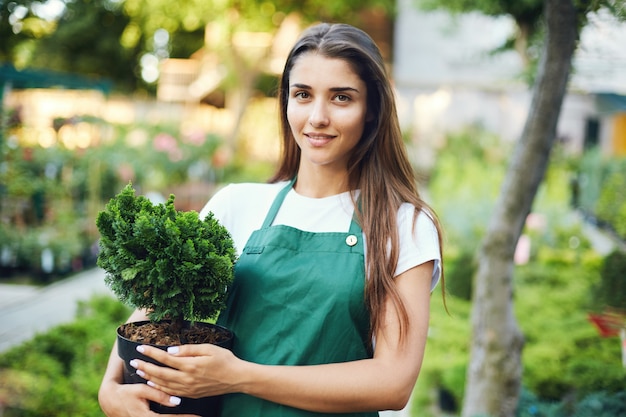 Female gardener holding a bonsai tree in a pot looking at camera. Owner of an outdoor garden supplies and plants store.