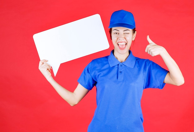 Female guide in blue uniform holding a white rectangular info board and showing enjoyment sign. 