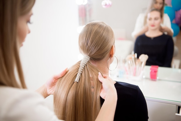 Female hairdresser makes a hairstyle to a blonde girl in a beauty salon