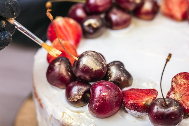 Female hand in black glove puts edible gold on cherries and strawberries on top of cheesecake with c