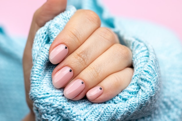 Female hand in a blue knitted sweater fabric with trendy beautiful manicure - pink nude nails with black small dots on a pink background. Selective focus. Closeup view