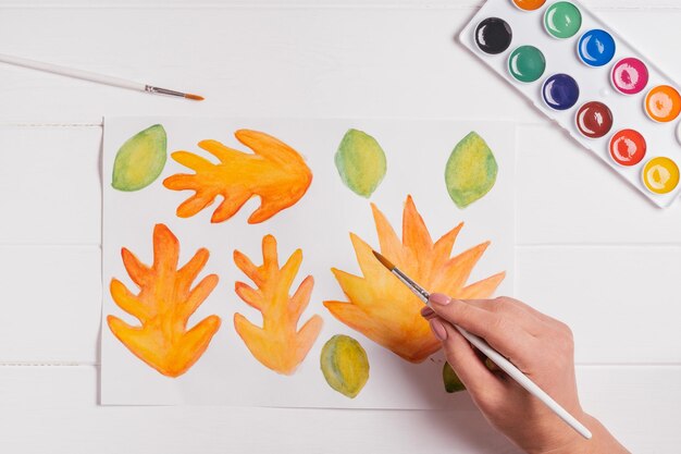 Female hand drawing autumn leaves different shapes sizes and colors with watercolors and brush on white table Top view