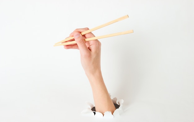 Female hand hold chopsticks through torn hole on white. Minimalistic food concept. Top view