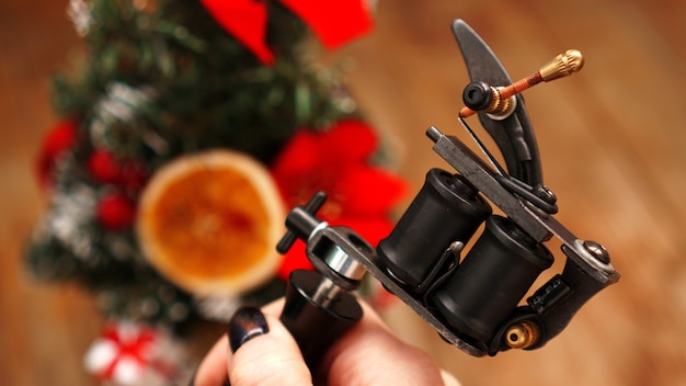 Female hand holding black tattoo machine on blurred christmas tree background