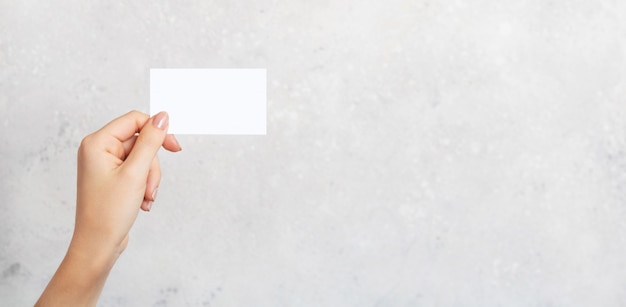 Female hand holding a blank business card, cutaway on gray concrete background with copy space. Branding mockup template.