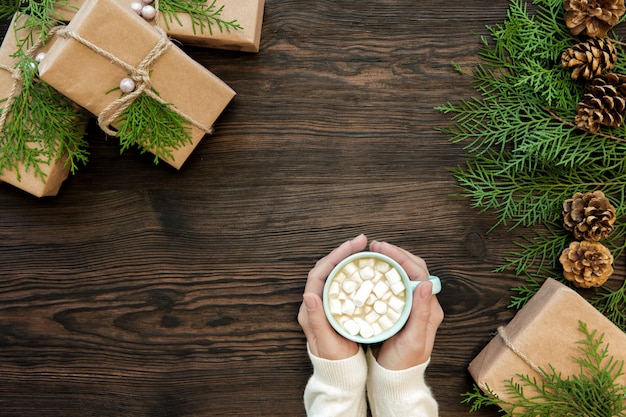 Female hand holding cup of chocolate with marshmallow and gift boxes on dark wooden, top view