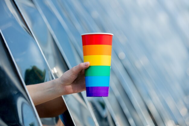 Female hand holding a cup in the color of the LGBT flag on the background of metal construction