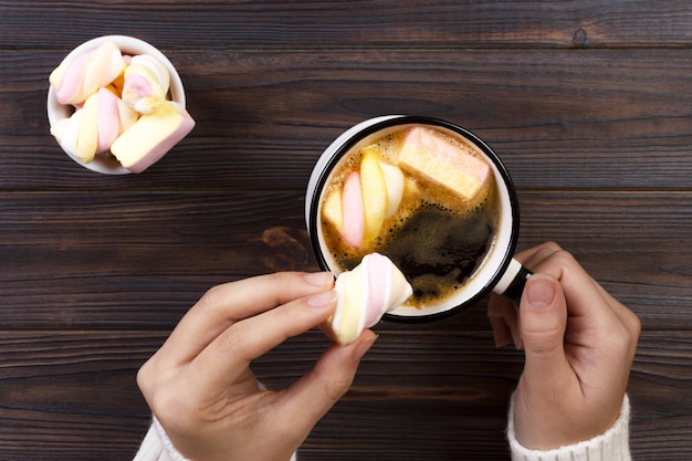 Female hand holding cup of hot cocoa or chocolate with marshmallow on wooden table from above