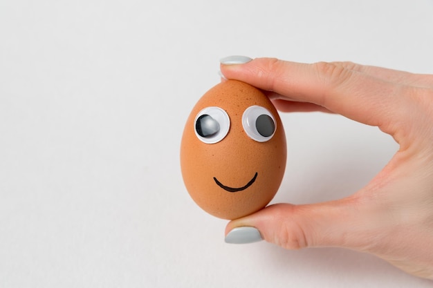 Female hand holding eggsmiley with googly eyes on white background Food with funny face