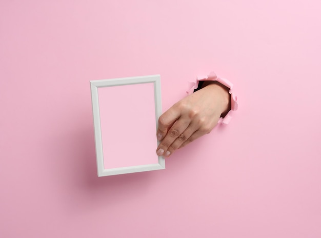 Female hand holding empty white wooden frame, body part sticking out of torn hole in pink paper background