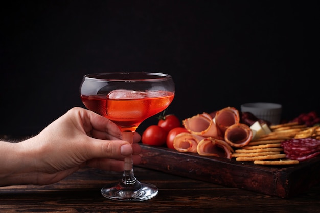 Female hand holding a glass of red liquor and charcuterie board, alcoholic cocktail with a appetizer, close-up.