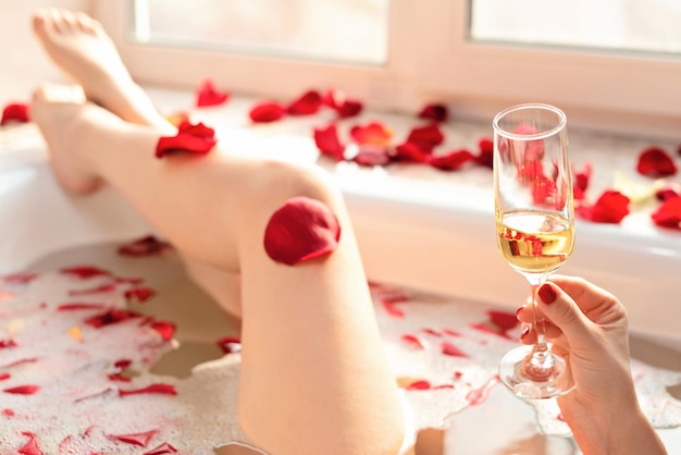 Female hand holding a glass of white wine while sitting in a bathtub with foam and red rose petals