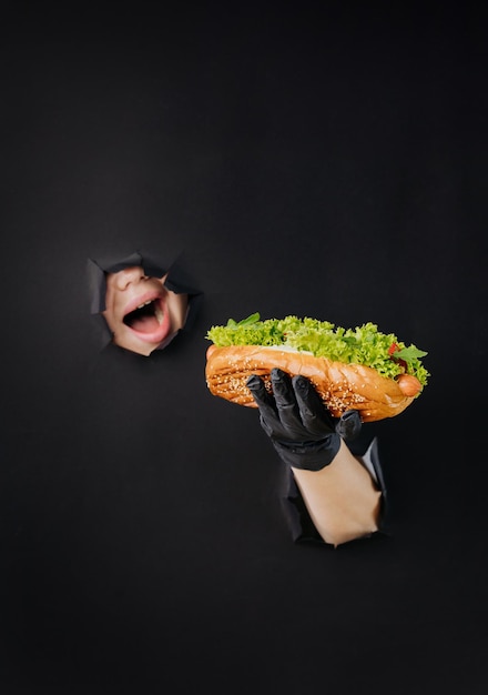 Female hand holding hot dog and looks through holes in black paper background street food concept