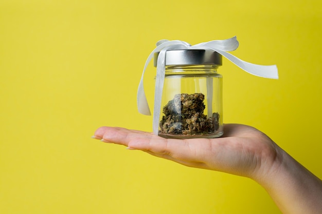 Female hand holding marijuana bud in glass jar with white ribbon on yellow background close up