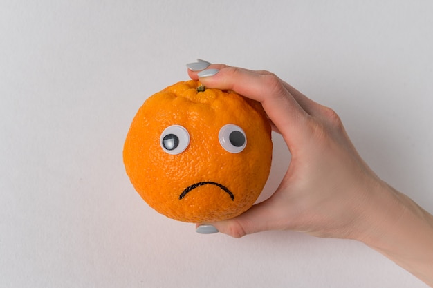 Female hand holding an orange-smile on white background. Orange with eyes and sad smile.