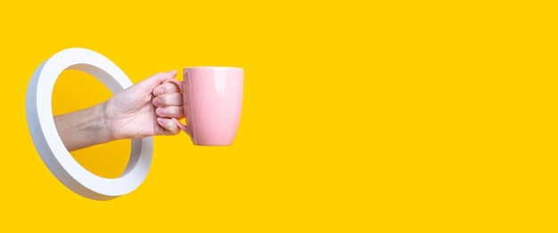 Female hand holding a pink cup in a round hole on a yellow background Banner