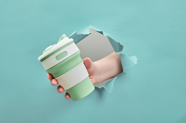 Photo female hand holding a silicone reusable cup through a hole torn. sustainable lifestyle. eco friendly concept.