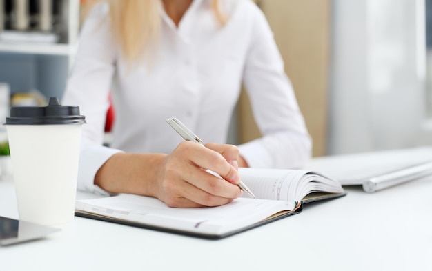 Female hand holding silver pen ready to make note in opened notebook sheet Businesswoman coffee cup workspace make thoughts records at personal organizer white letter collar conference signature