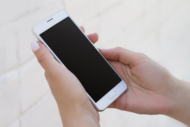 Female hand holding smartphone on white brick wall background, mockup with copy space