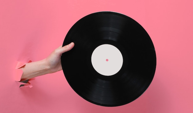 Female hand holding vinyl record through the torn pink paper background. Minimalistic retro concept