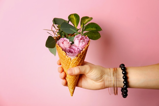 Female hand holding waffle cone with composition of flowers and branches on pink background