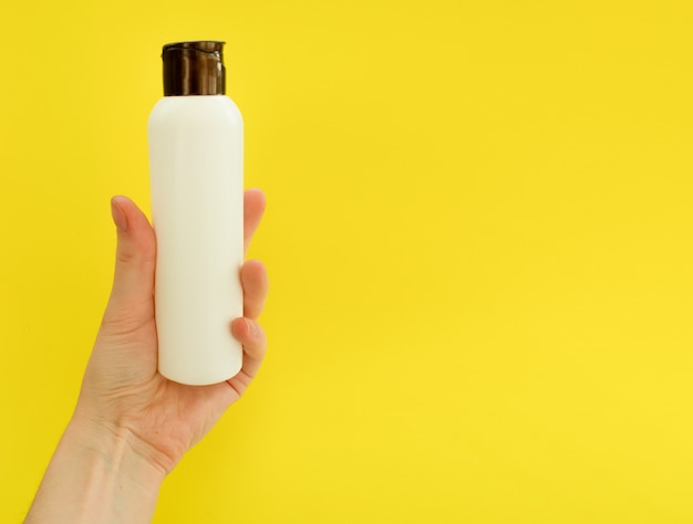 Female hand holding white cosmetic bottle on yellow background mock up copy space summer skincare co...