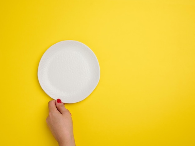 Female hand holding a white empty round plate on a yellow background top view copy space