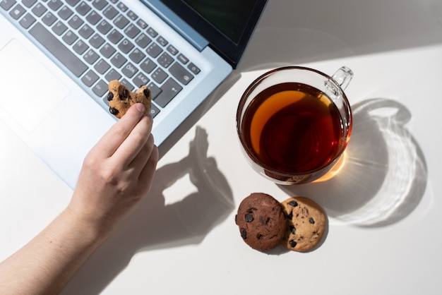 Photo a female hand holds a bitten chocolate chip cookie next to a laptop, a cup of tea, two cookies in sunlight, top view.