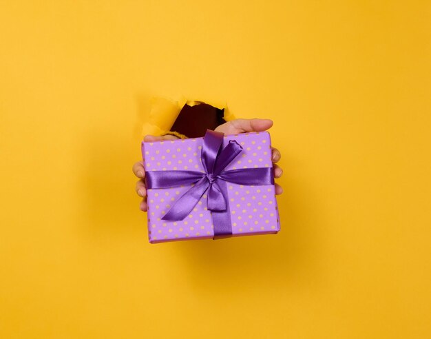 Female hand holds a box with a gift on a yellow background, part of the body sticks out of a torn hole in a paper background. Congratulation, holiday surprise