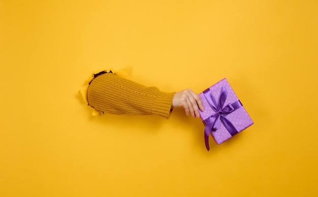 Female hand holds a box with a gift on a yellow background, part of the body sticks out of a torn hole in a paper background. Congratulation, holiday surprise