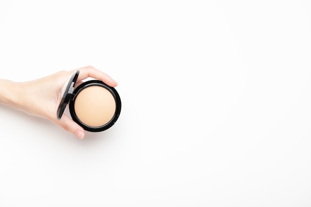 A female hand holds a compact beige powder on a white background, top view. Makeup nude product for perfect skin tone. Cosmetic, concealer, skin corrector. Copy space.