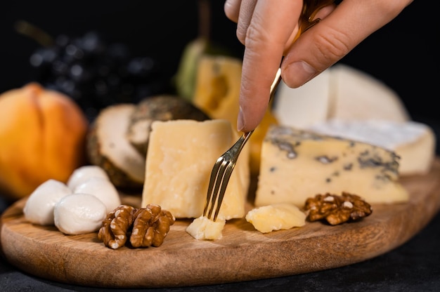 A female hand holds a dessert fork with parmesan. Nearby are mozzarella, gorgonzola, camembert. A combination of cheese and fruit. Close-up.