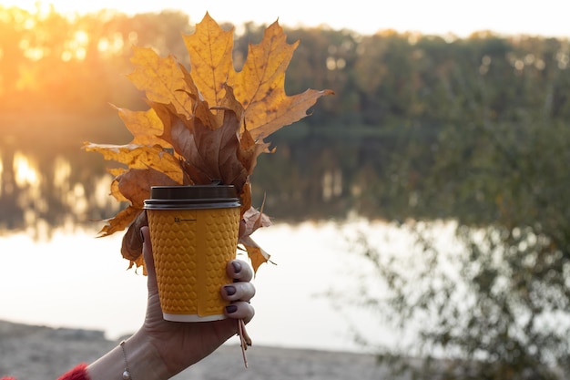 Female hand holds a glass of coffee with autumn leaves on the background of the river