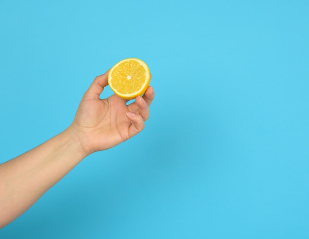 Female hand holds half a yellow lemon on a blue background, copy space