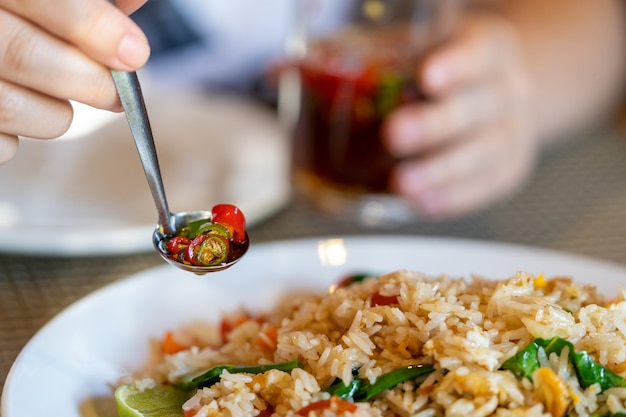 Female hand holds a metal spoon with seasoning of red and green pepper, close-up, on the background of rice and a glass.