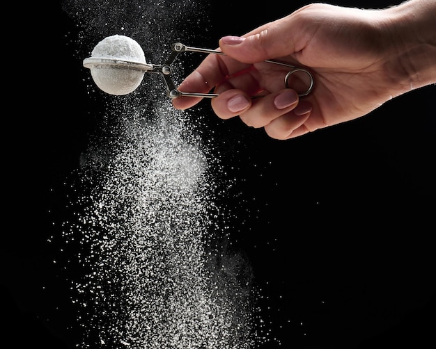 Female hand holds a metal strainer with powdered sugar Particles fly in different directions on a black background sprinkling