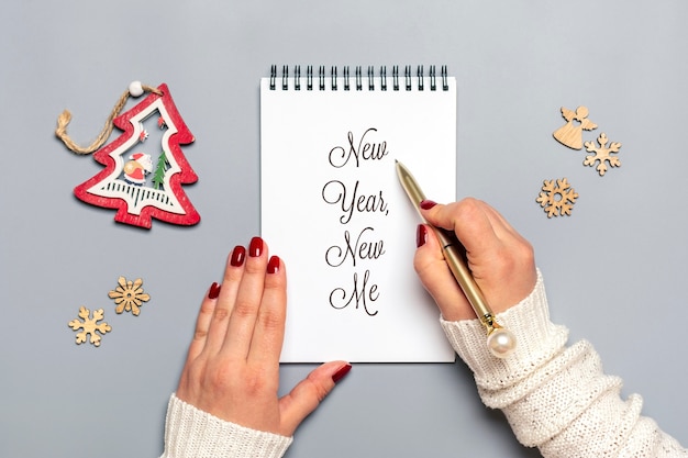 Female hand holds pen and writes New Year, New Me on white notepad, tree, snowflake on gray