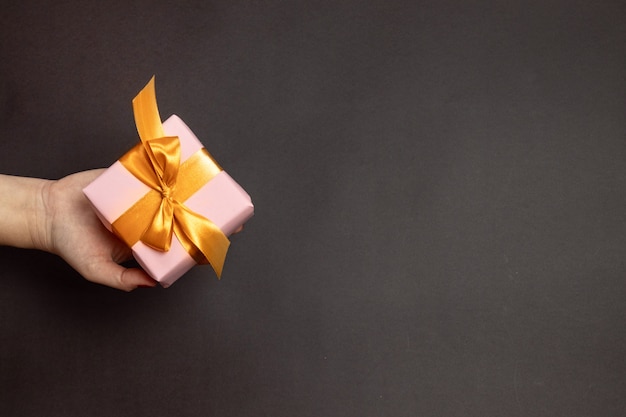 Female hand holds a surprise gift wrapped in a pink box and a gold ribbon on dark