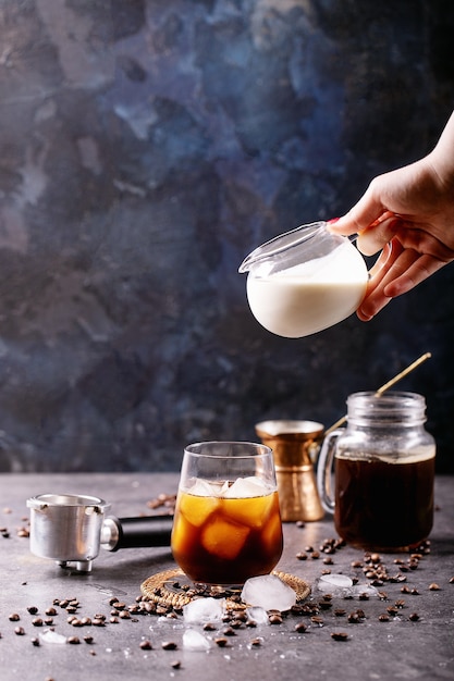 Female hand pouring cream into ice coffee over blue wall with coffee beans and ice cubes laying around