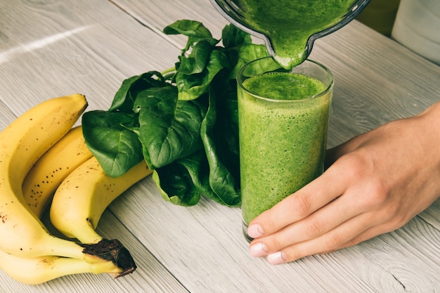 Female hand pours a smoothie of banana and spinach in glass on a wooden background