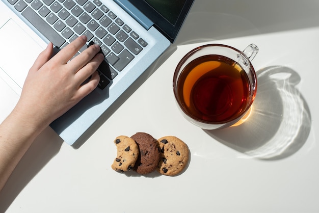 Photo female hand prints on a laptop, a cup with tea, and three chocolate chip cookies in sunlight, top view.