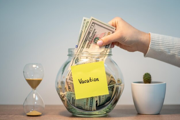 Female hand putting stack of dollars in glass jar with inscription Vacation