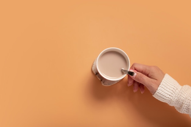 Female hand in sweater holding cup of cocoa on colored background top view with copy space