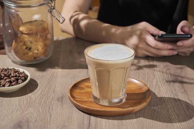 Female hand used and play phone waiting drink Young woman in cafe or restaurant drink americano or hot latte coffee from glass in wooden table with cookie in jar breakfast beverage morning concept