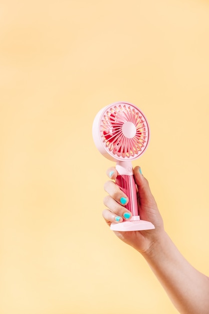 Female hand with blue nail polish holding a pink electric fan on yellow background